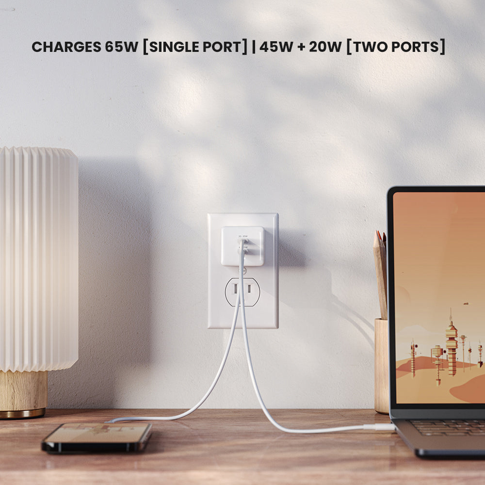 Laptop and phone charging on a wooden desk with a lamp and wall outlet in the background.